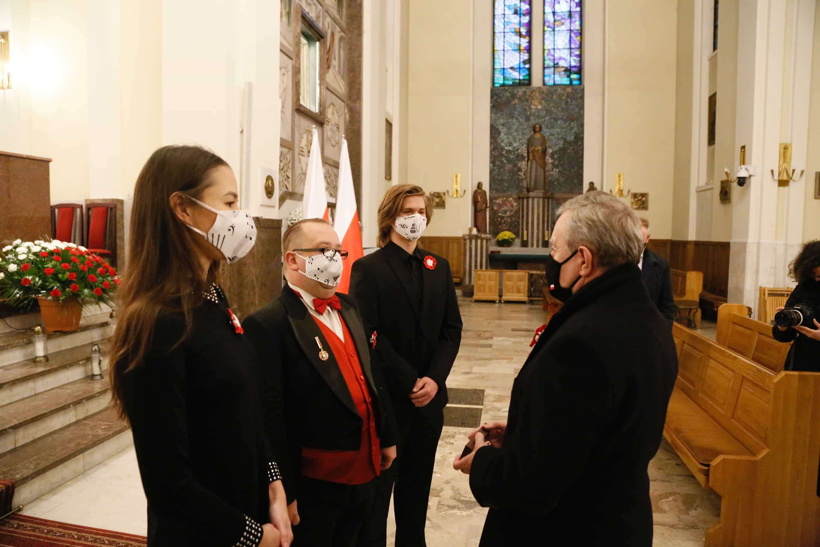 Kościół katolicki. W centrum trzech mężczyzn i jedna kobieta. Mężczyzna po prawej stronie nosi poprawnie założoną na nos i usta czarną maseczkę, a osoby po lewej poprawnie założone białe maseczki z nutami