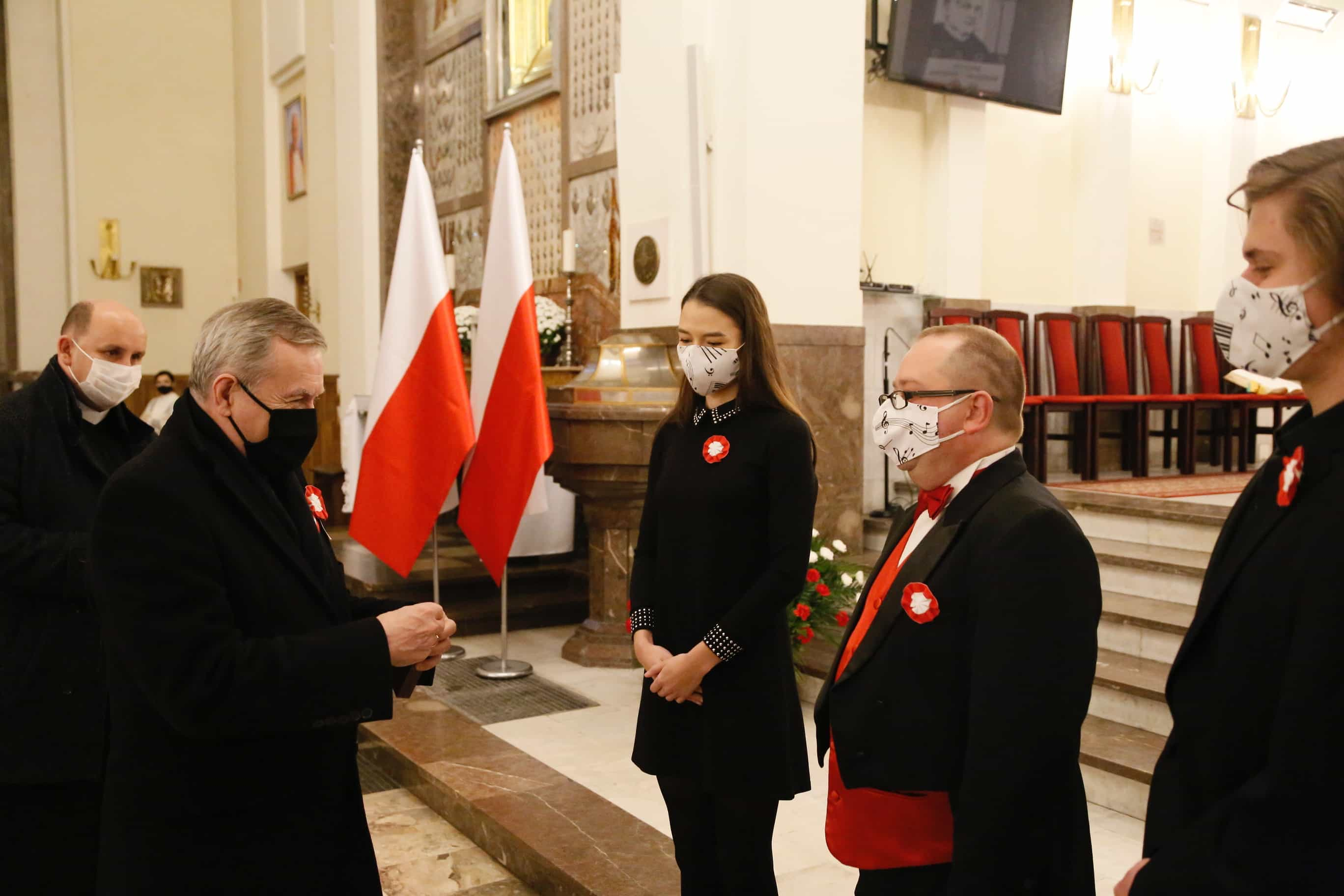 zdjęcie przedstawiające pięć osób ubranych na czarno. Cztery z nich to mężczyźni, a w centrum widoczna jest kobieta. W tle są dwie flagi Polski, telewizor, czerwone krzesła z brązowymi nogami wykonanymi z drewna oraz marmurowa podłoga